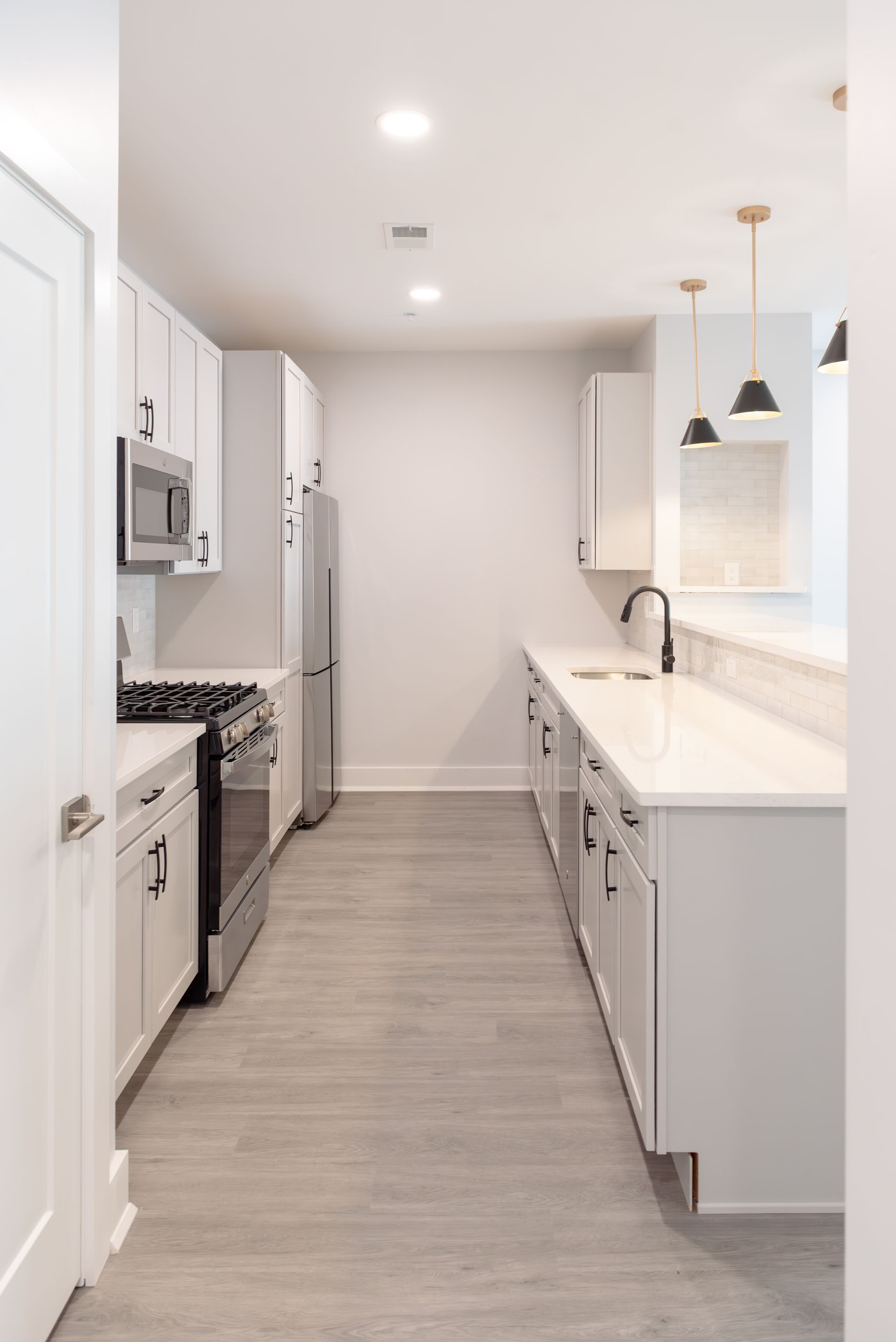 Narrow, white kitchen with stainless steel appliances and light wood-look flooring.