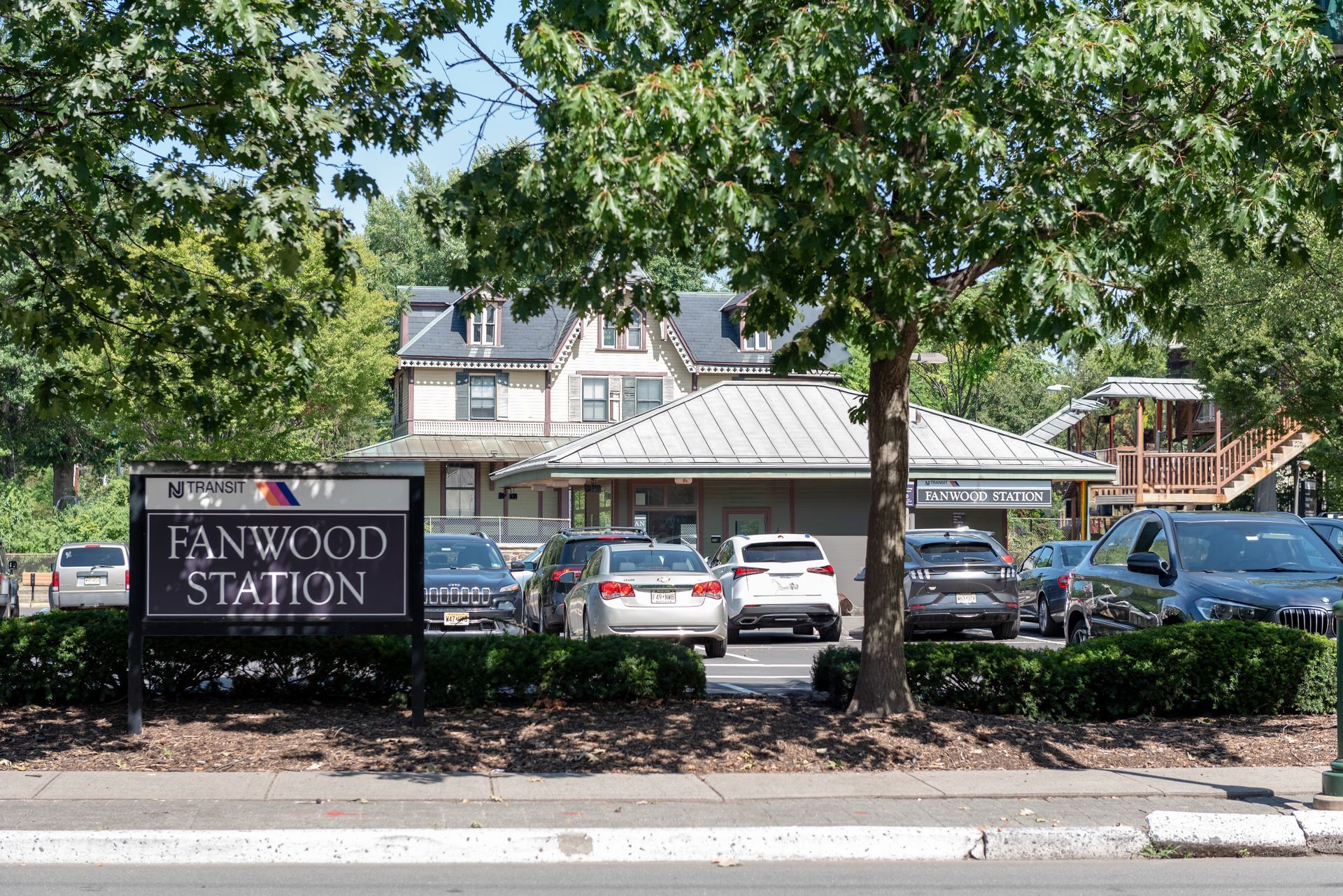 Fanwood Station sign in front of a building and parked cars, under a bright sunny sky.