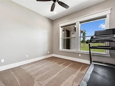 Empty room with gray walls, tan carpet, treadmill by a window, and ceiling fan.