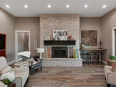 Living room with brick fireplace, hardwood floors, neutral colors, and various furniture.