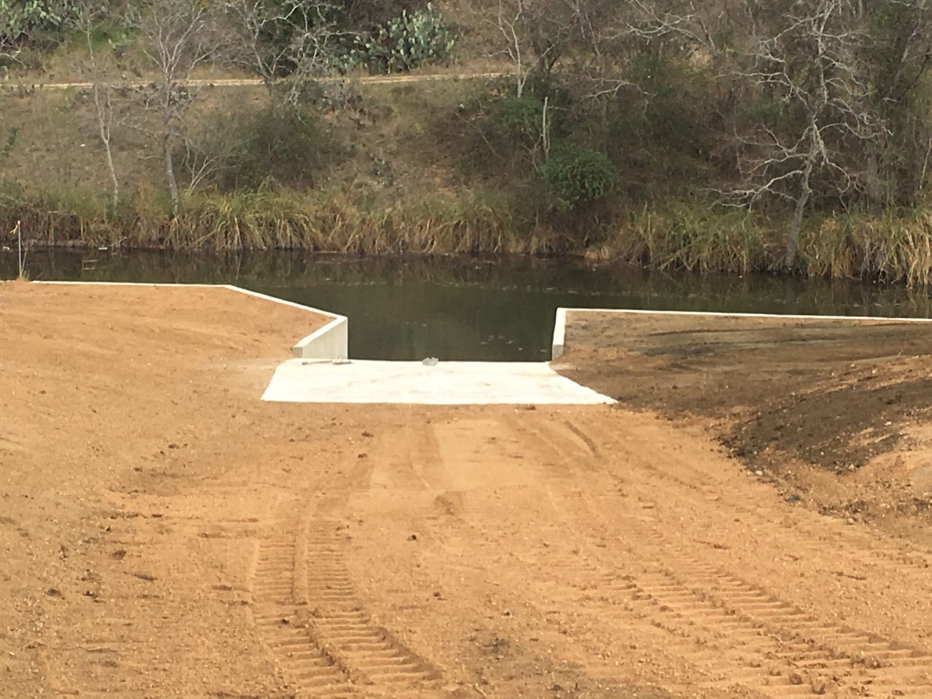 Boat ramp leading into a dark body of water, surrounded by dirt and trees. Excavation project.