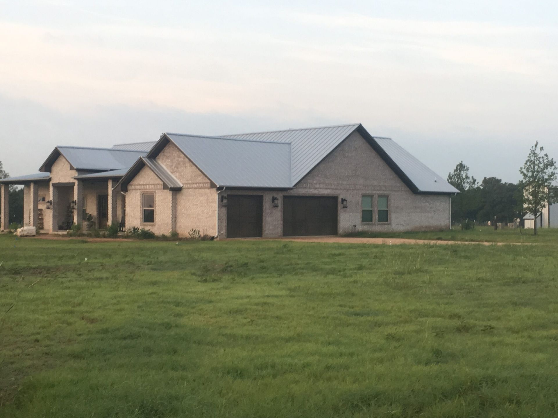 A brick house with a gray metal roof and two-car garage sits in a grassy field.