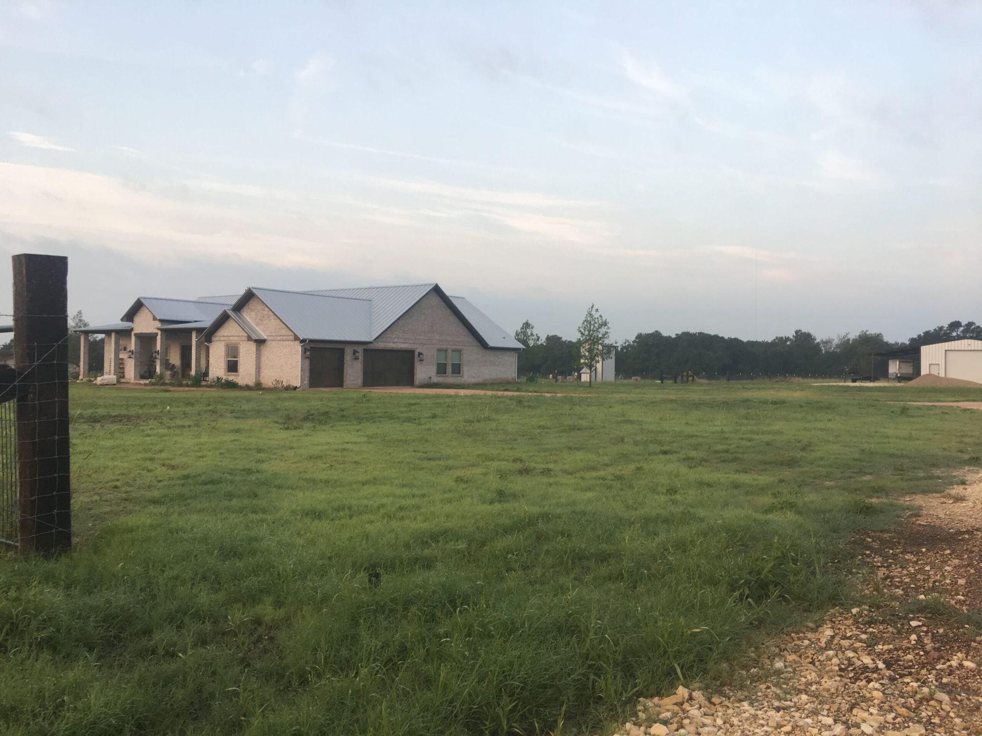 A single-story house with a metal roof, surrounded by green grass and a dirt road.