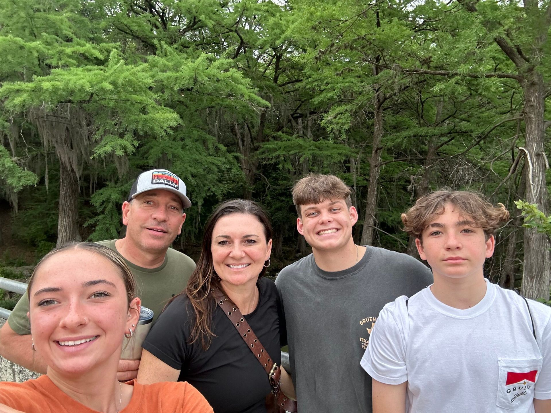 Family smiling outdoors; trees in the background.