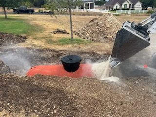 A backhoe fills a trench with water, burying a septic tank with a black lid. Outdoors.