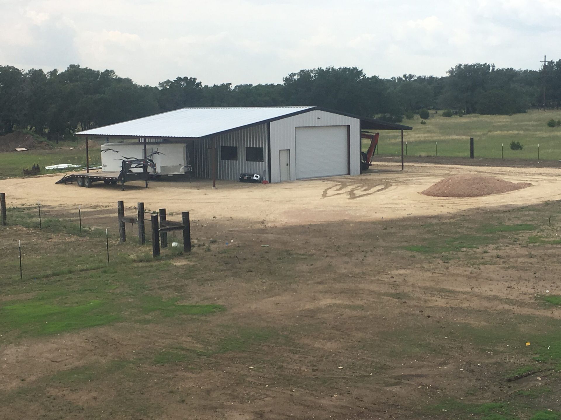 A metal-roofed building with an open front on a dirt lot in a rural setting, with trees in the background.