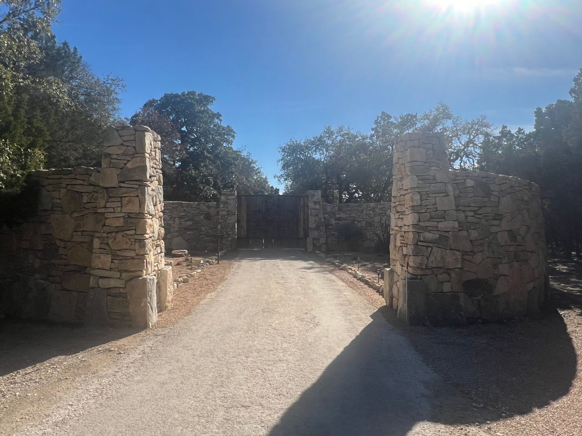 Stone entrance pillars frame a dirt road leading to a wooden gate on a sunny day.