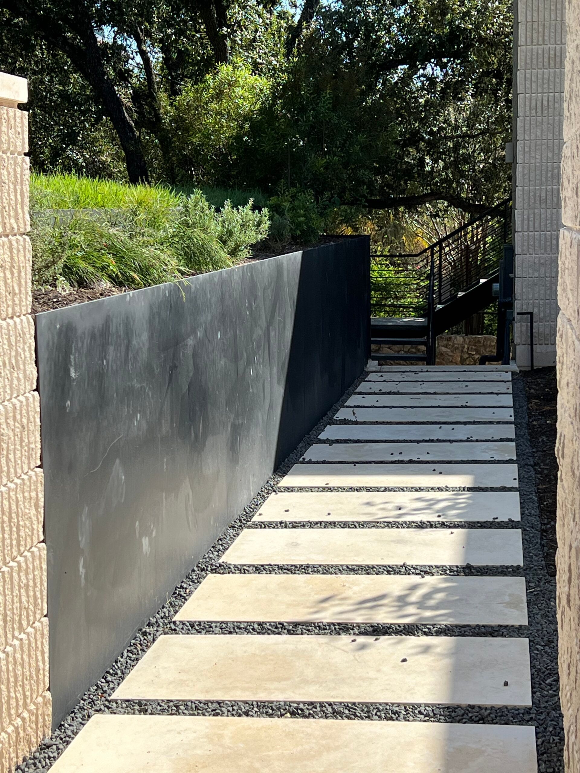 Concrete pathway with a dark wall, leading to stairs in a sunny, outdoor setting.