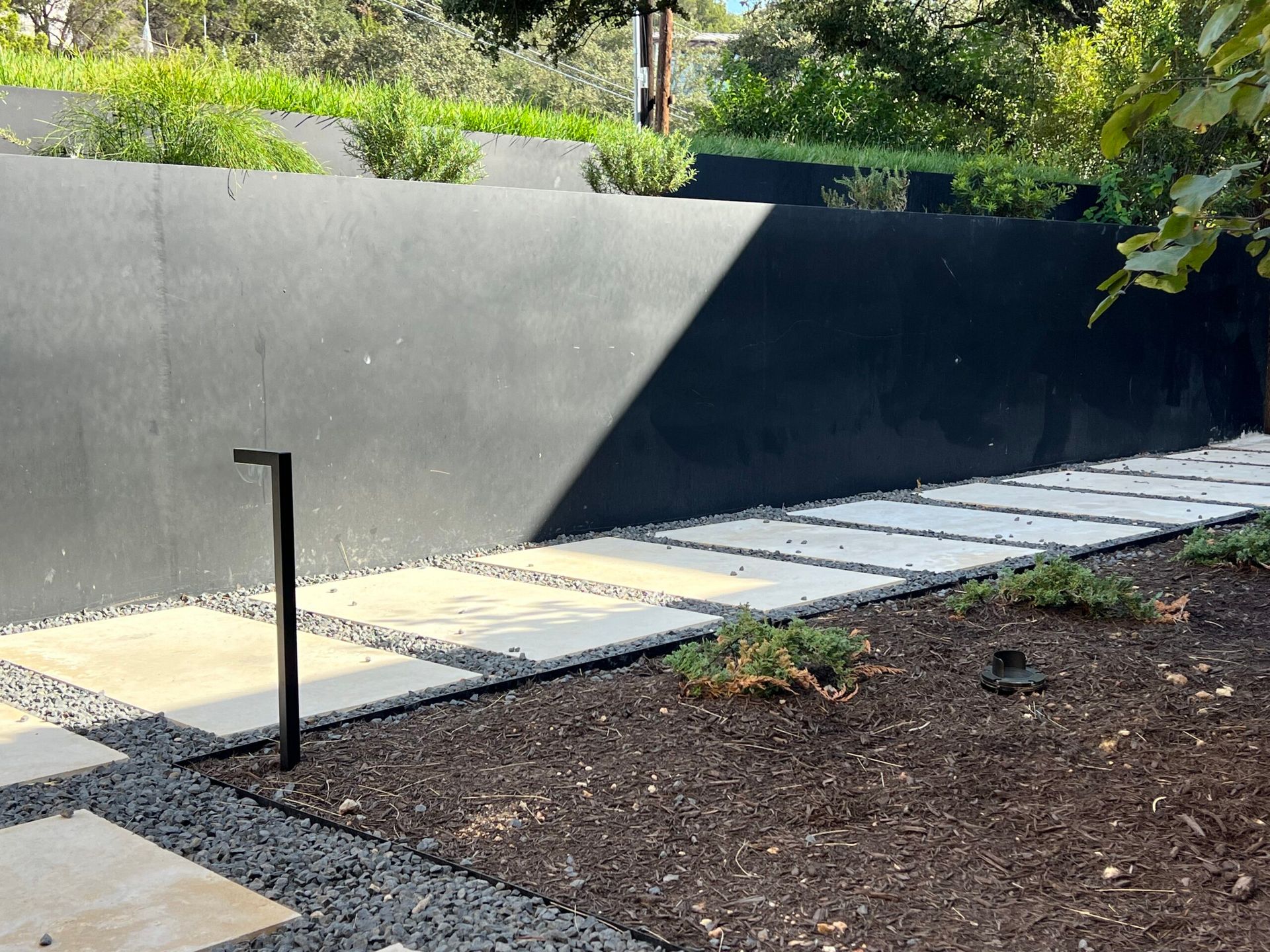 Stone path along black wall, with garden, grass, and modern light fixture.
