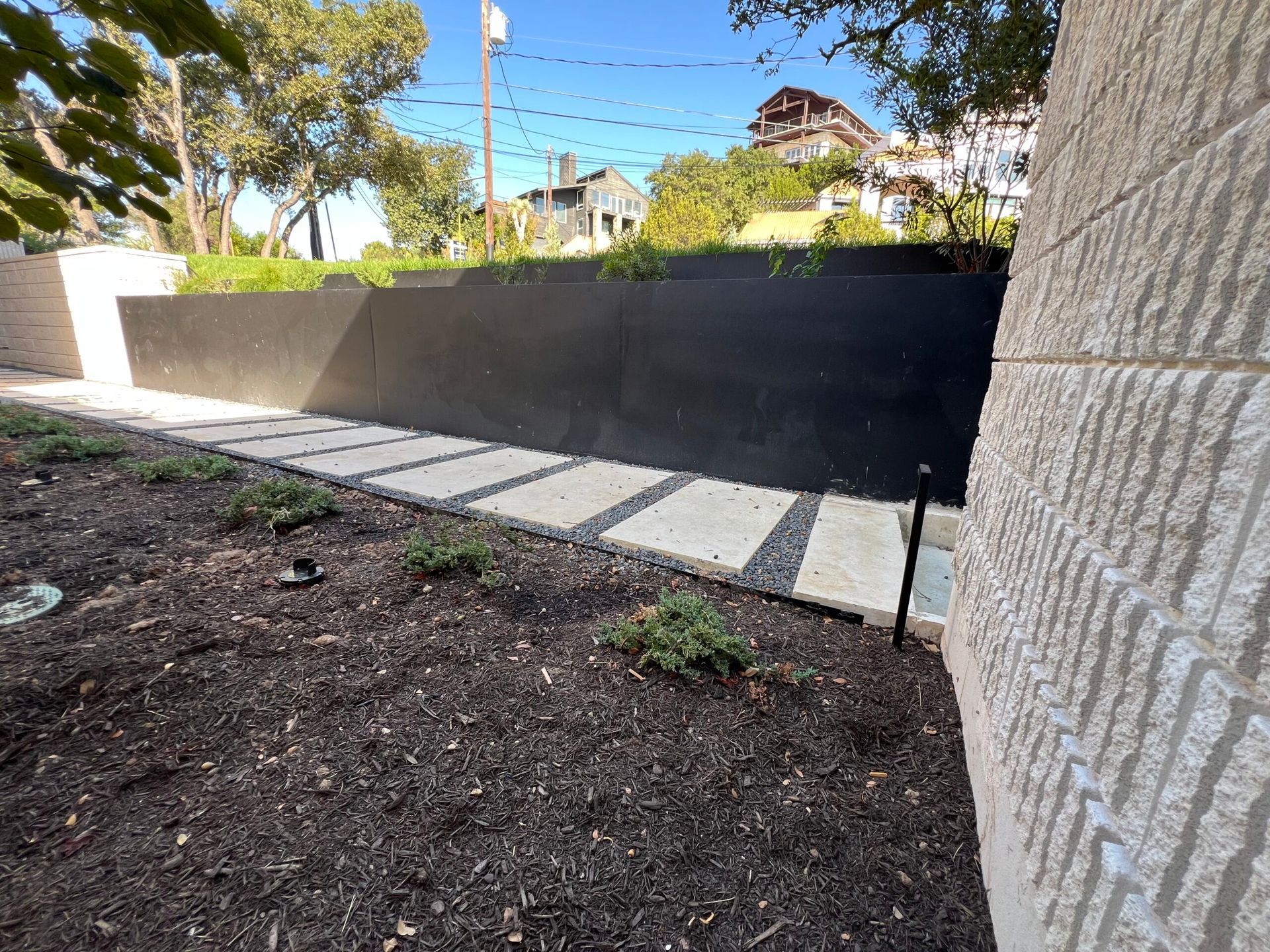 Stone walkway alongside a black wall and textured stone building in a landscaped yard.
