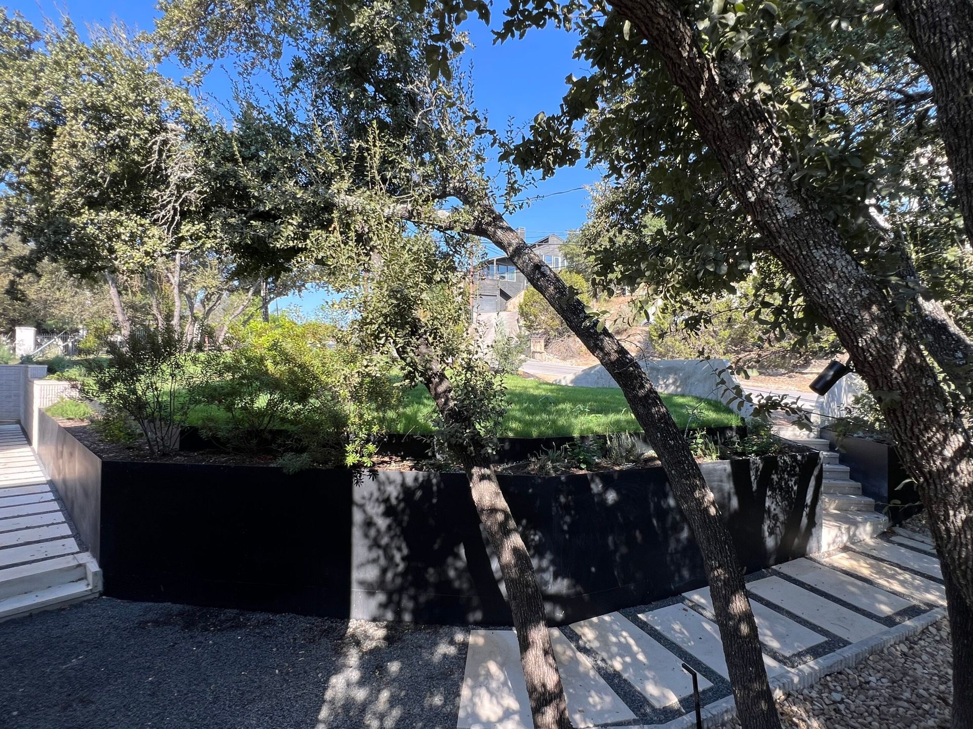 Landscape with trees, green plants, black retaining wall, and a pathway under a bright blue sky.