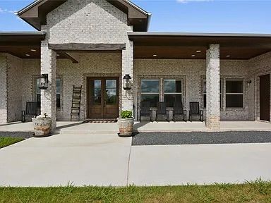 A brick home with a porch, pillars, and wooden door, with potted plants and chairs.