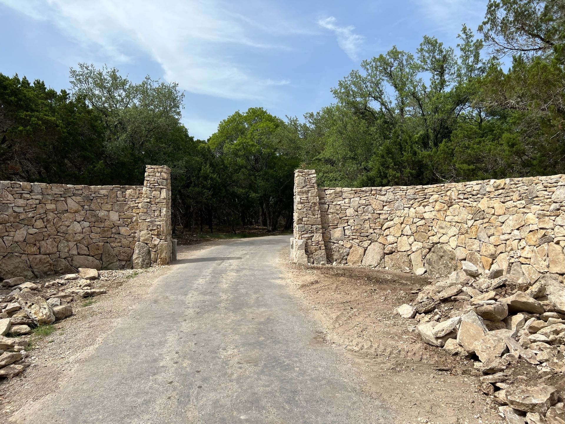 Stone walls frame a gravel road leading into a forest under a blue sky.