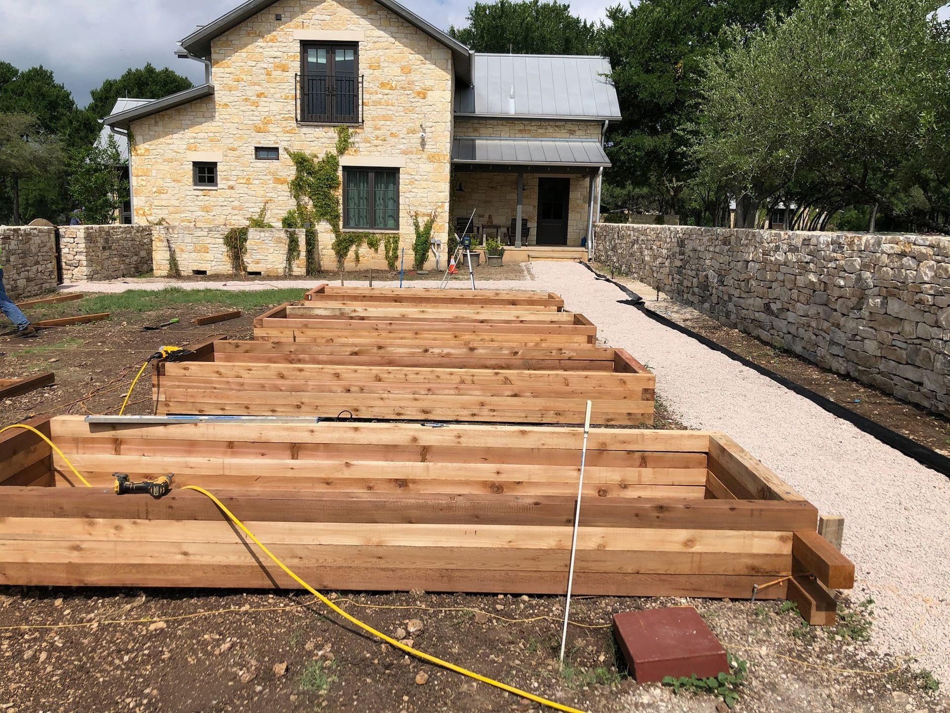 Raised wooden garden beds in front of a stone house with a gravel path and stone wall.