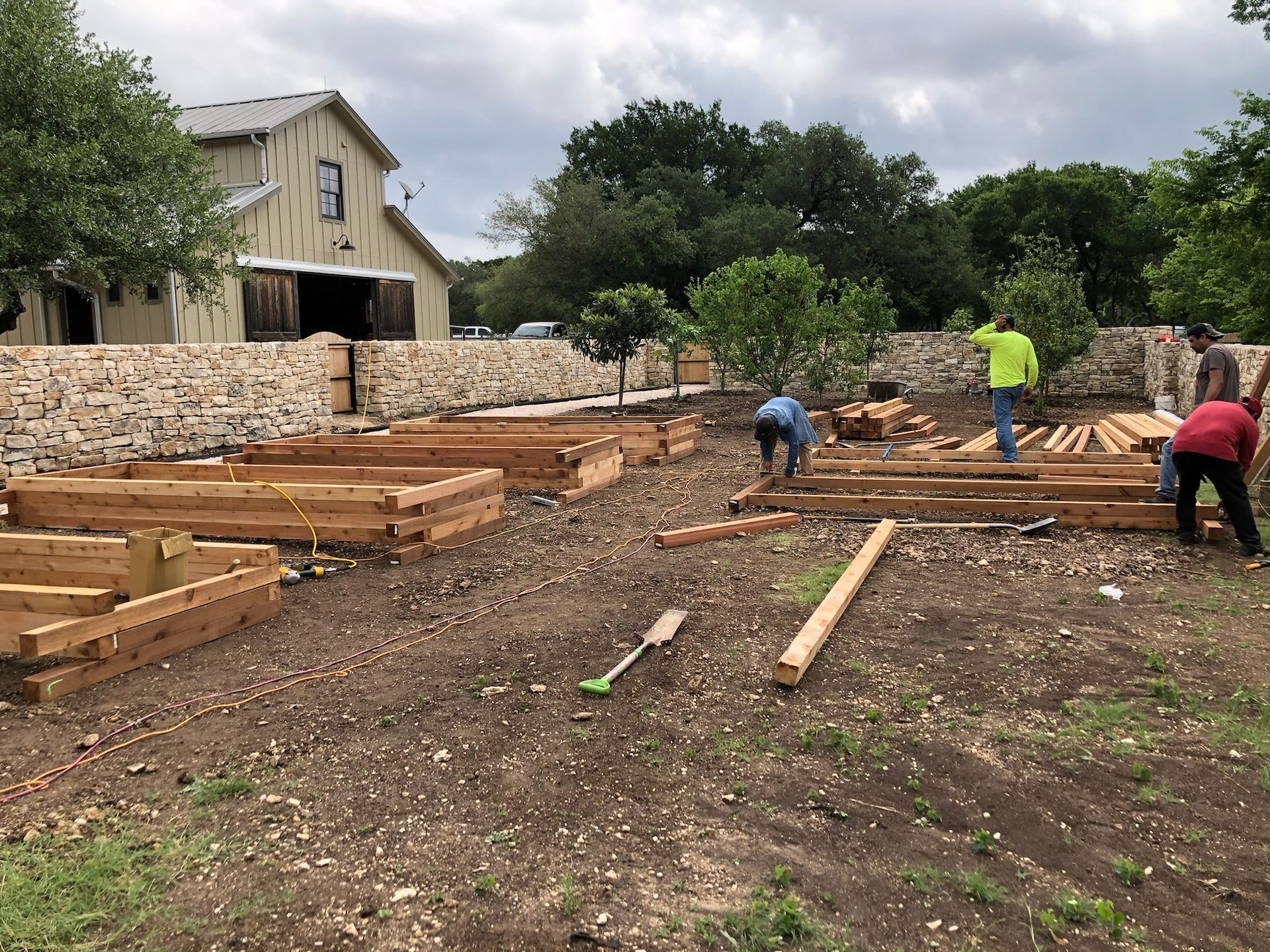 Men constructing wooden garden beds on a brown, dirt plot with a stone wall and barn in the background.