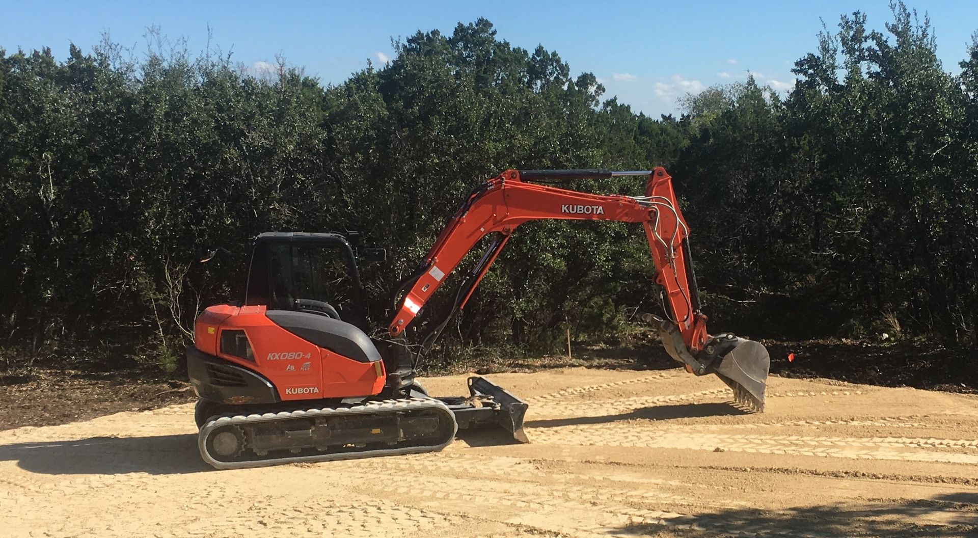 Orange Kubota excavator digging on a sandy surface, trees in background.