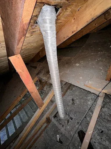 Corrugated white vent pipe rising through an attic floor beside wooden beams and a metal duct.