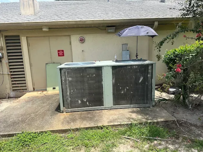 Outdoor HVAC unit beside a house wall, with a patio umbrella and flower bushes nearby