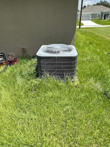 Outdoor air conditioner unit beside a house wall on a grassy lawn; bicycle partly visible in the corner.