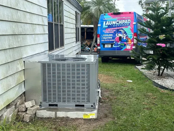 Outdoor HVAC unit beside a house with a colorful LaunchPad van parked on the grass nearby