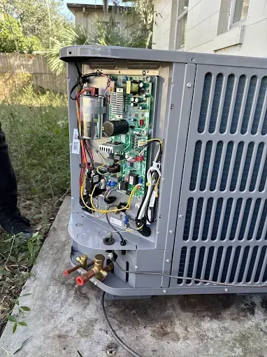 Outdoor HVAC unit with front panel removed, exposing wiring and components on a concrete pad.
