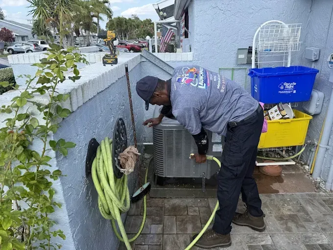 Person bending over an outdoor AC unit, rinsing it with a hose beside a house and cleaning supplies.