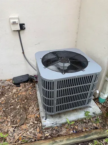 Outdoor air conditioner unit beside a wall, with a fan on top and fallen leaves around it