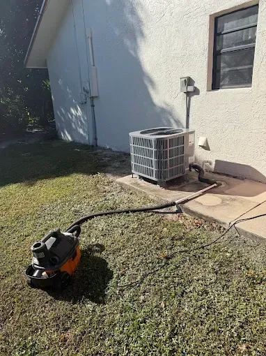 A small lawn mower on grass beside a house wall and outdoor air conditioner unit.