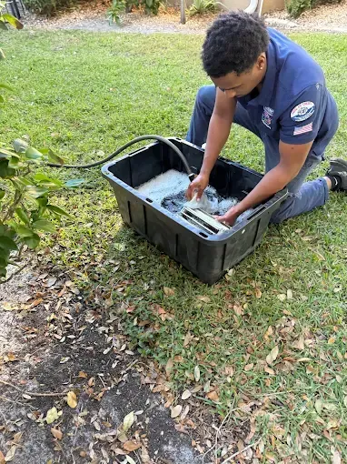 Person kneeling outdoors, washing an item in a black plastic tub on the grass and pavement.
