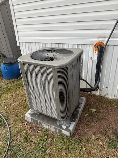 Outdoor air conditioner unit beside a house wall, with a blue barrel and hose nearby