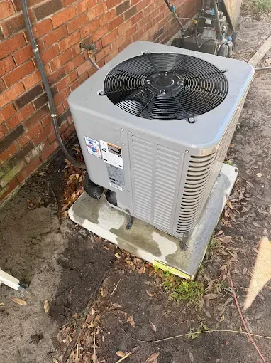 Outdoor air conditioner unit beside a brick wall, with a fan on top and water stains on the base.
