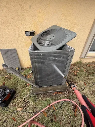 Outdoor HVAC condenser unit with top fan exposed, beside a house wall and yard tools on the ground