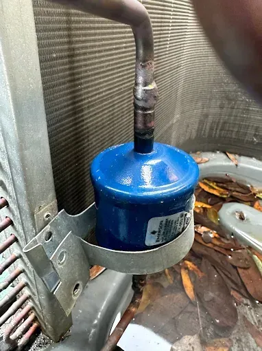 Blue cylindrical HVAC component with a copper pipe, mounted in a metal housing with leaves below
