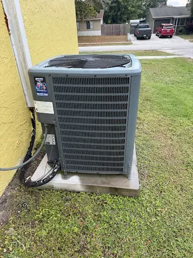 Outdoor air conditioning unit beside a yellow house wall on a concrete pad, with grass and driveway nearby