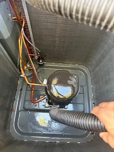 Hand holding a vacuum hose inside a metal HVAC unit, cleaning around a black fan or pump assembly.