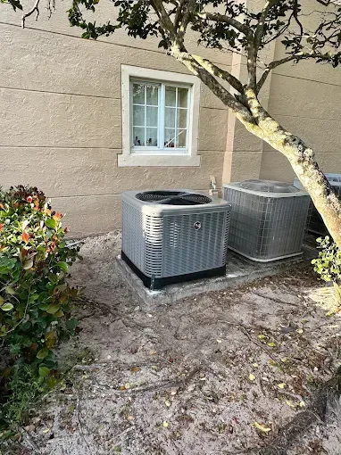 Two outdoor air conditioner units beside a beige wall under a tree, with a small window above.