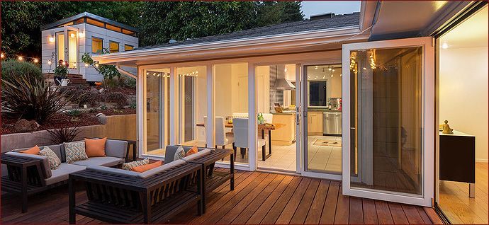 Outdoor patio with a seating area, opening to a kitchen, and a separate building on a hillside.