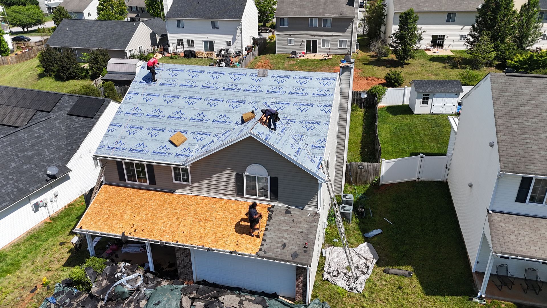 Roofers working on a house with partial new roofing. The sky is blue, and the yard is green.
