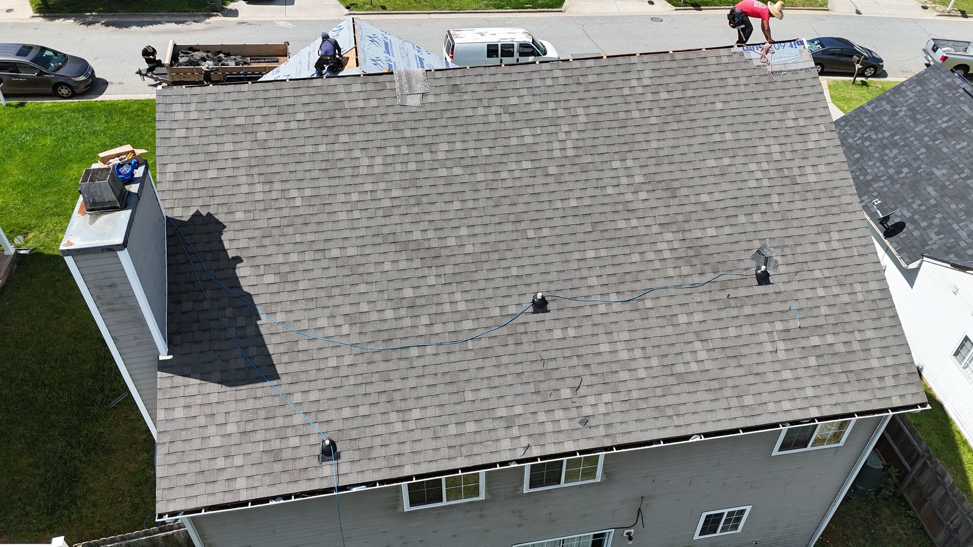 Roofers working on a gray shingled roof of a two-story house on a sunny day.