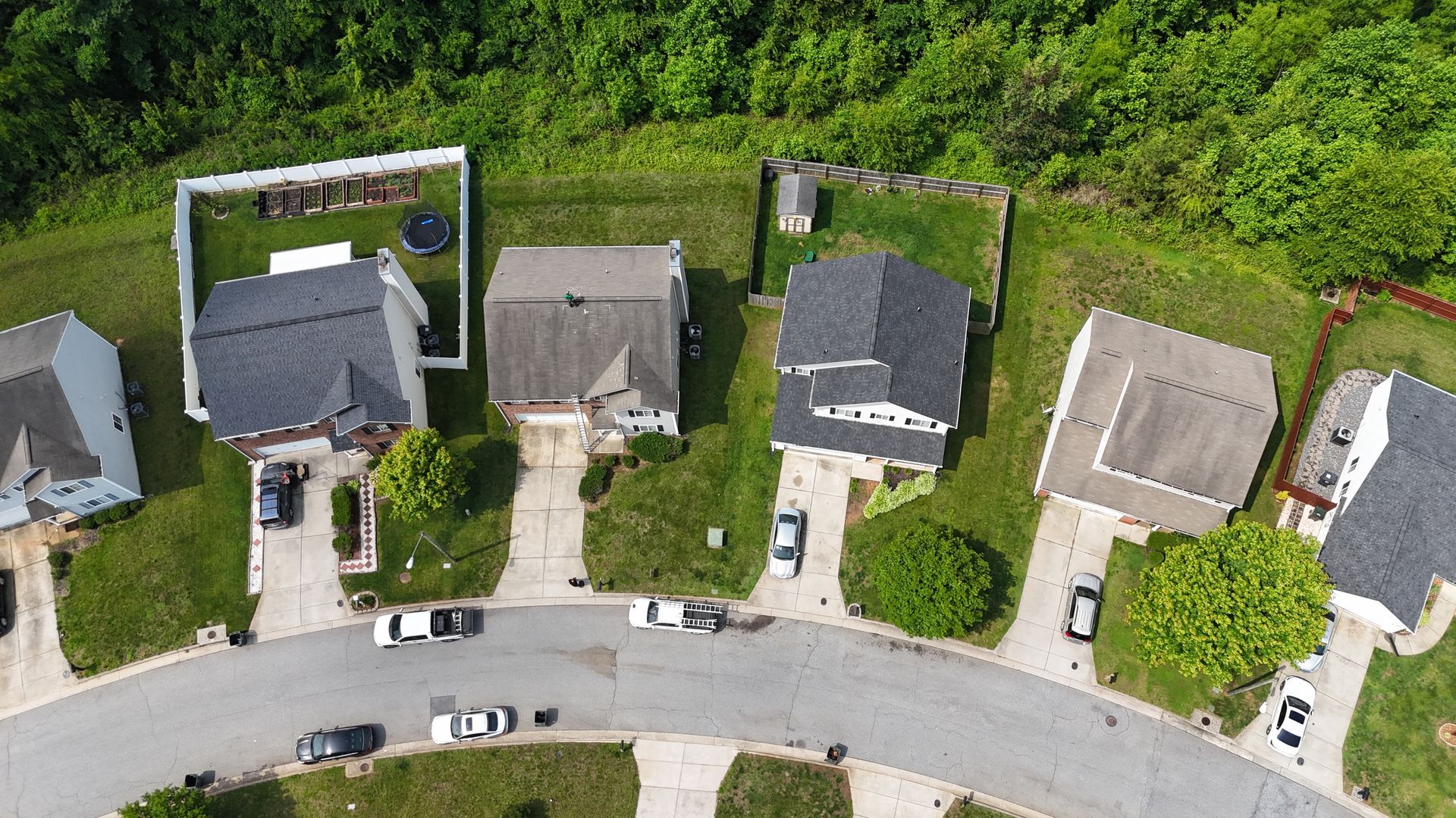 Aerial view of suburban houses with cars parked on the street and driveways, surrounded by green lawns and trees.