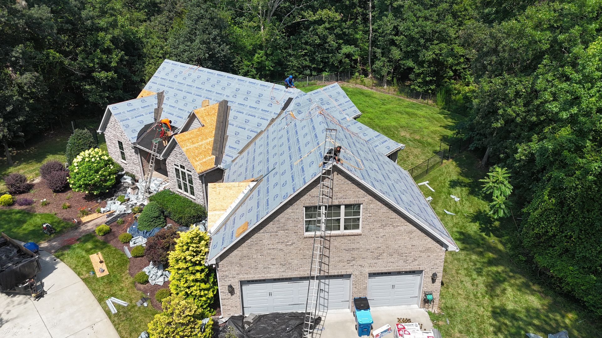 Overhead view of a house with a partially installed gray roof, surrounded by trees and greenery. Two car garage visible.