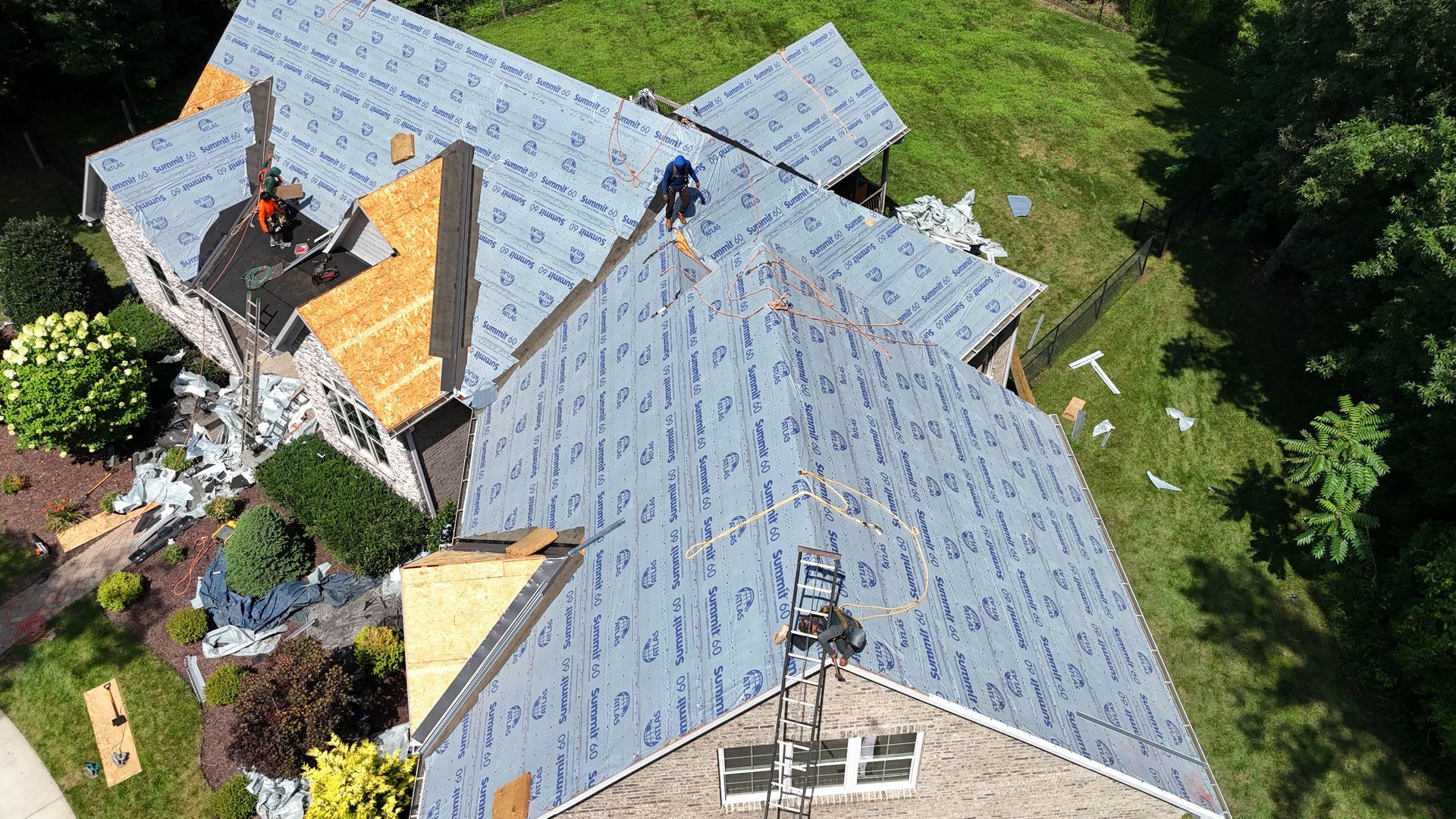 Roofers working on a multi-gabled roof, covered with blue underlayment. Green trees and yard surround the house.