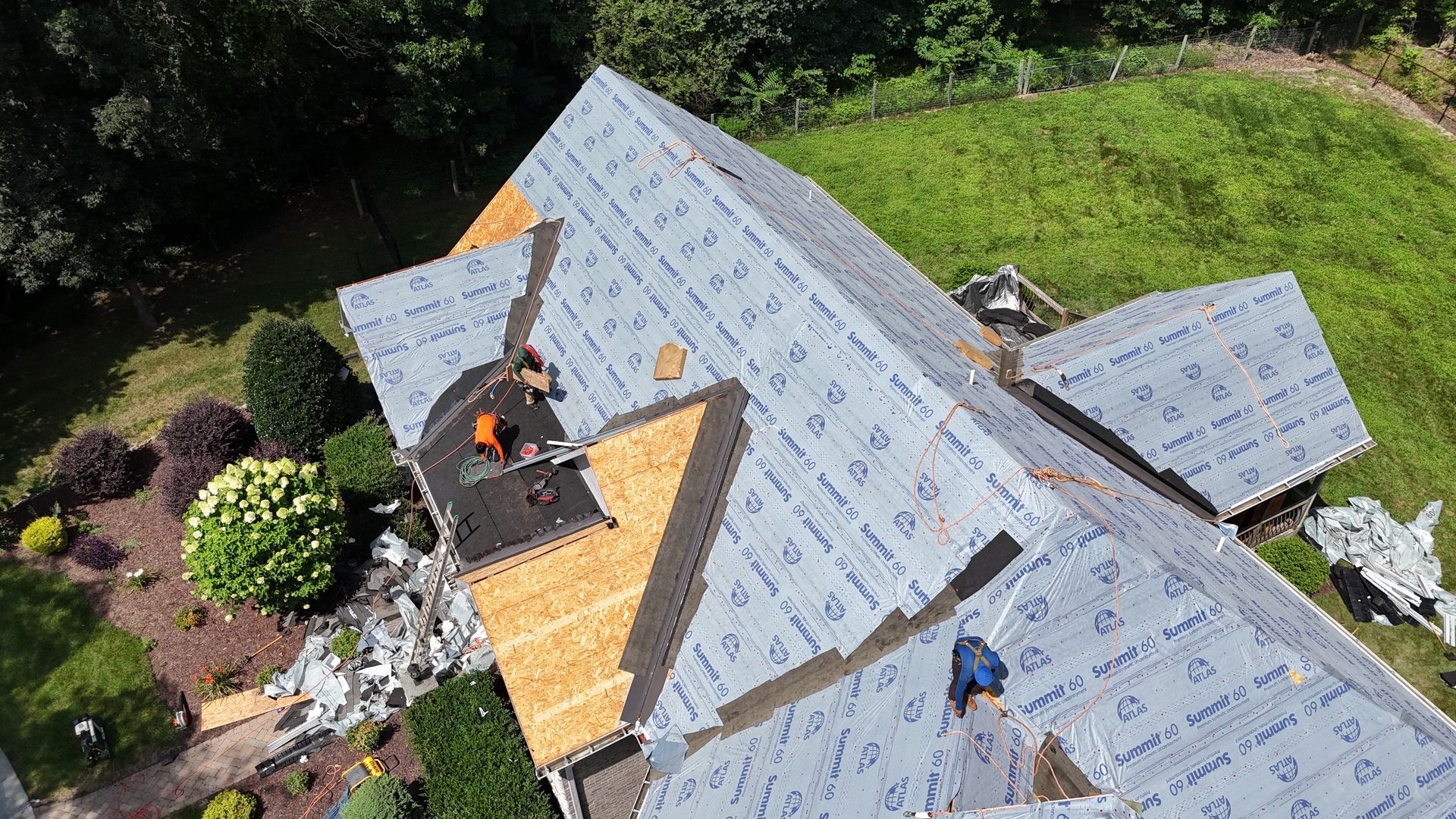 Roofers working on a house roof. Shingles being installed on a partially covered roof. Green lawn and shrubs surround the house.