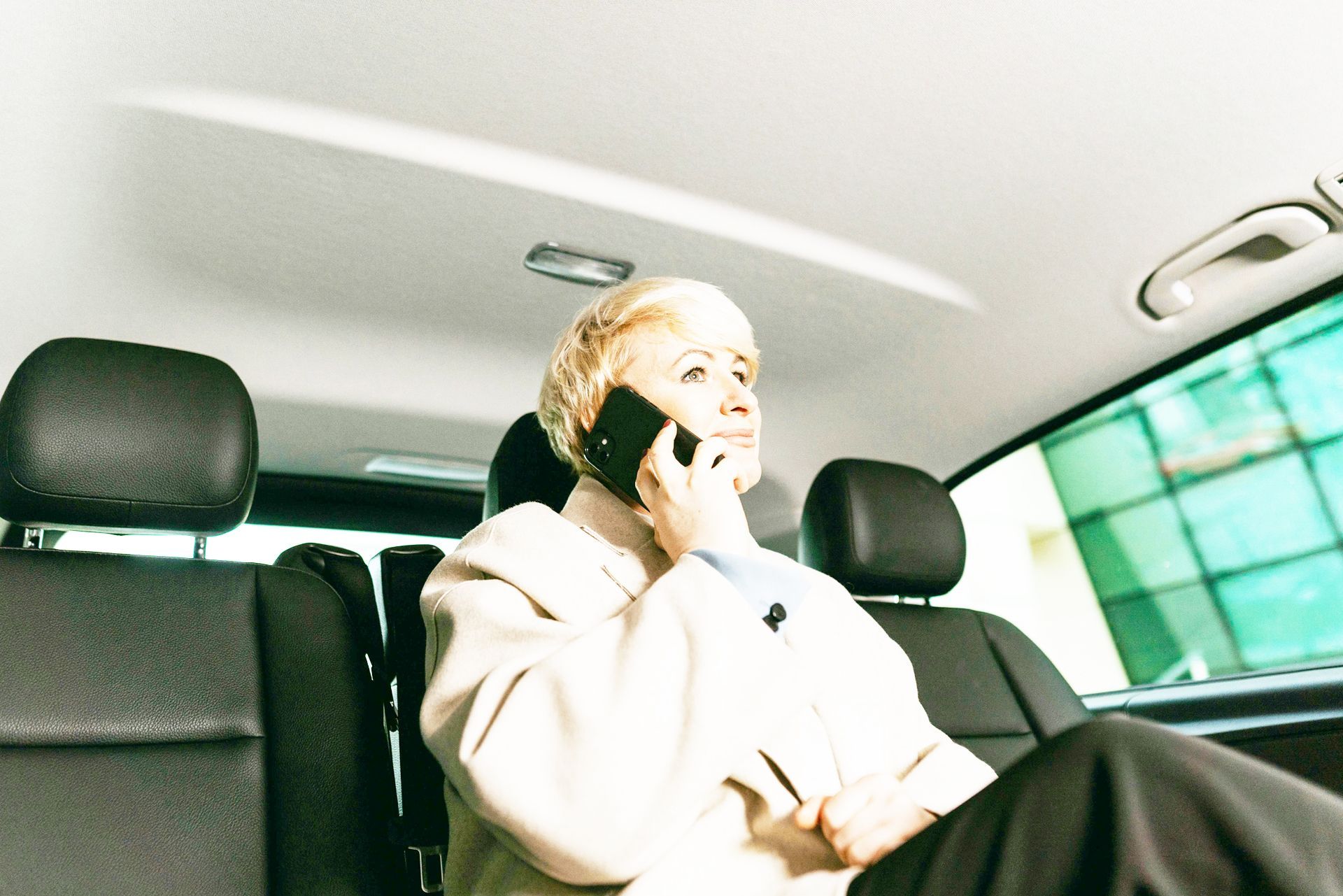 Woman in a car talking on a cell phone, wearing a beige coat, black van seats.