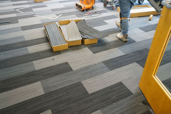 A worker installs gray and black modular carpet tiles in an office space, with some tiles still in cardboard boxes.