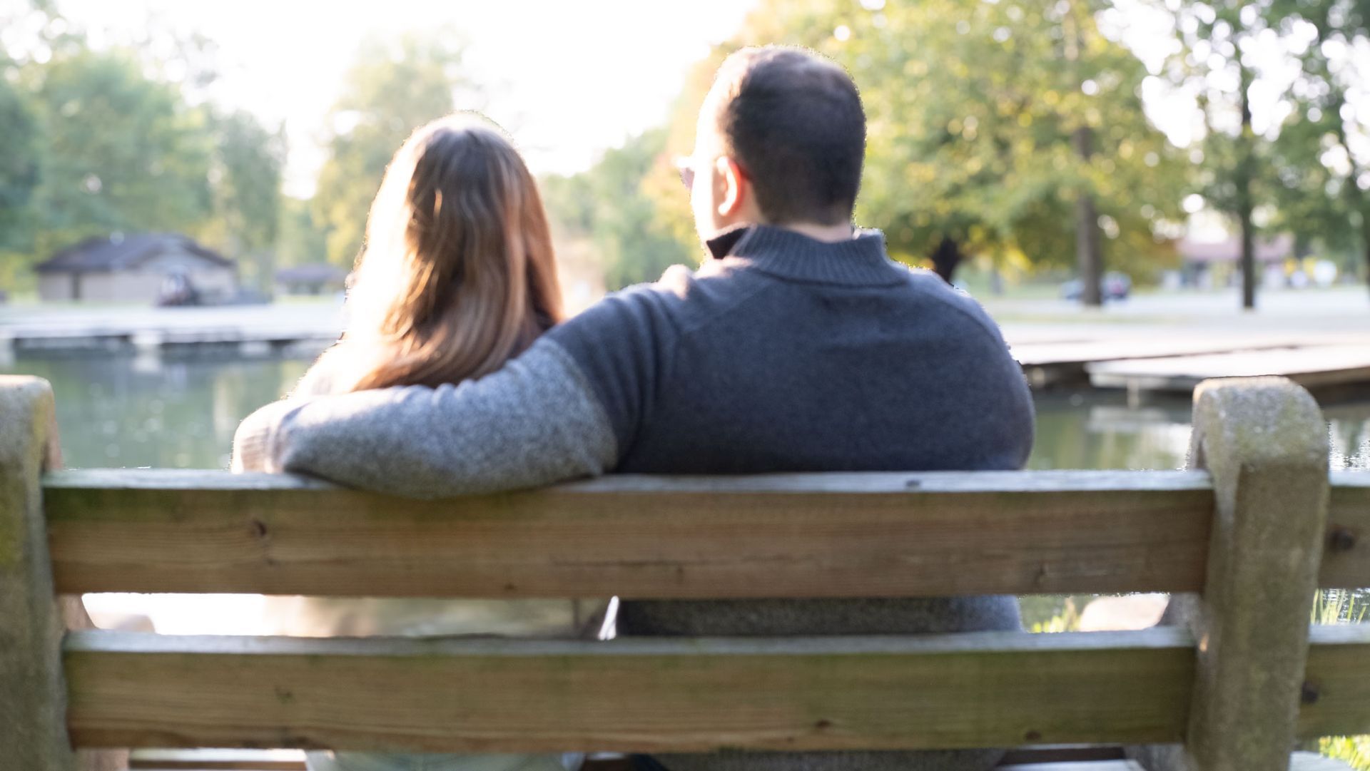 Two people sitting on a wooden bench by a pond, looking at the water in a sunny park.