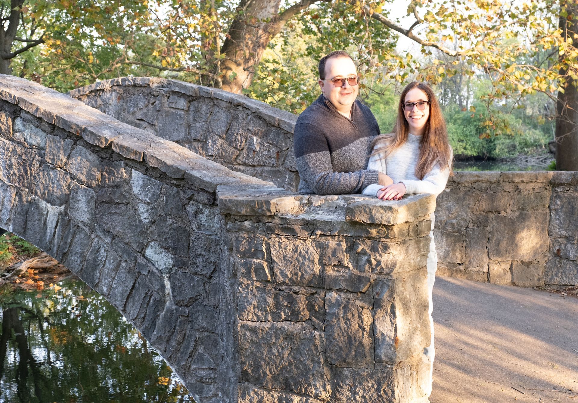 Two people leaning on a stone bridge in a wooded park, wearing sunglasses and looking at the camera.