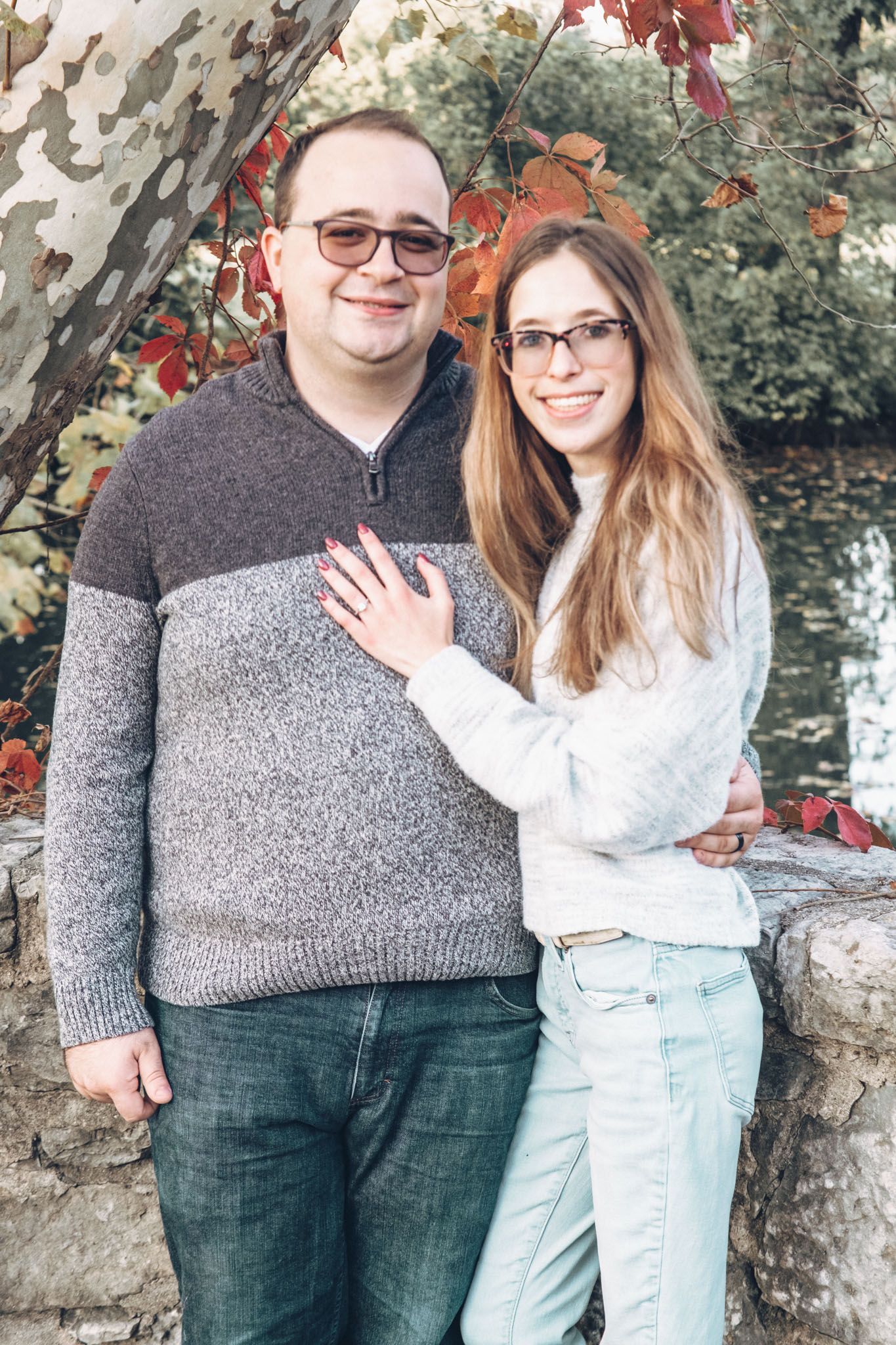 Two people standing together outdoors, smiling beside a stone wall and autumn foliage.
