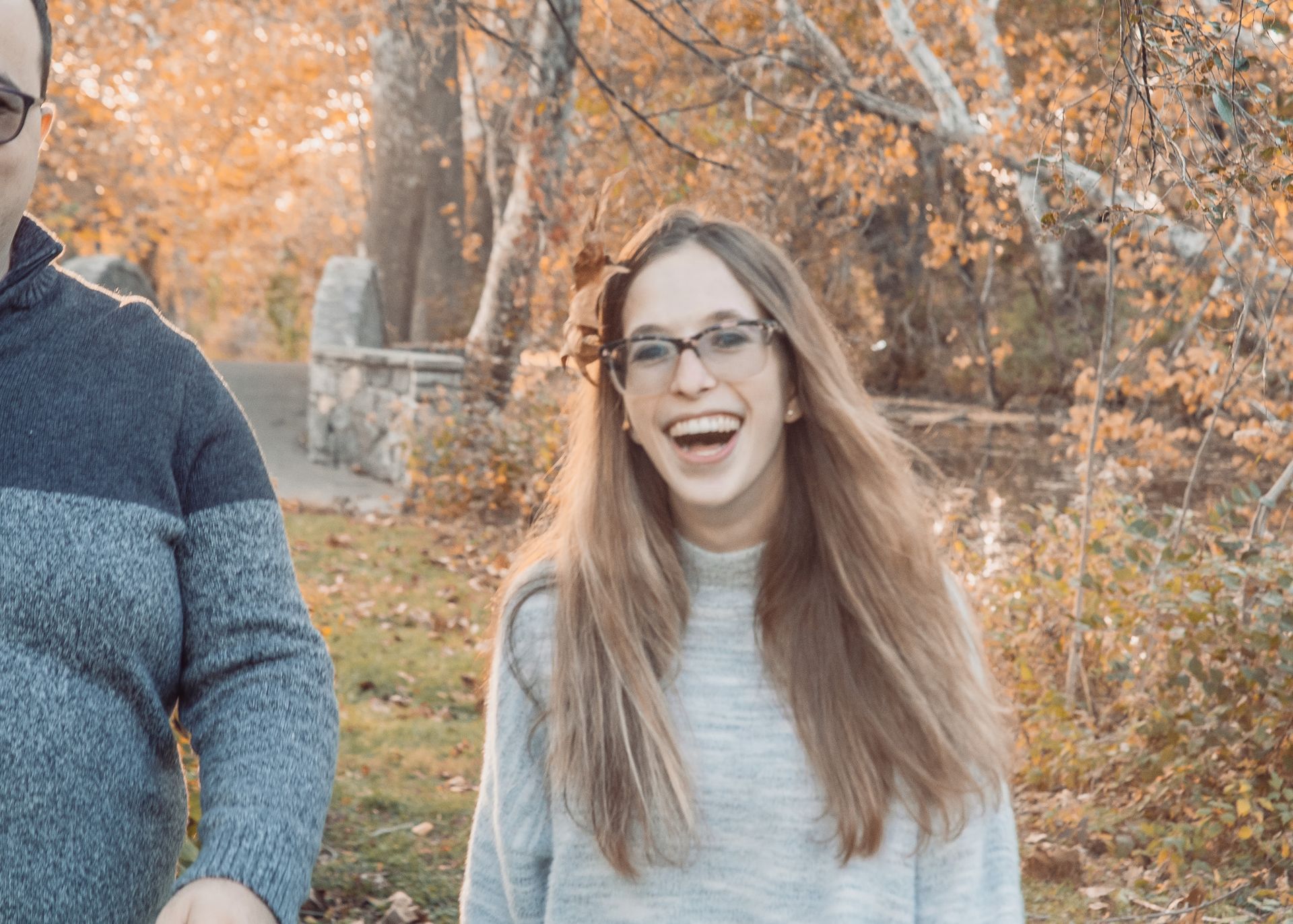 Smiling woman with long hair and glasses standing outdoors in autumn, with a person partly visible beside her