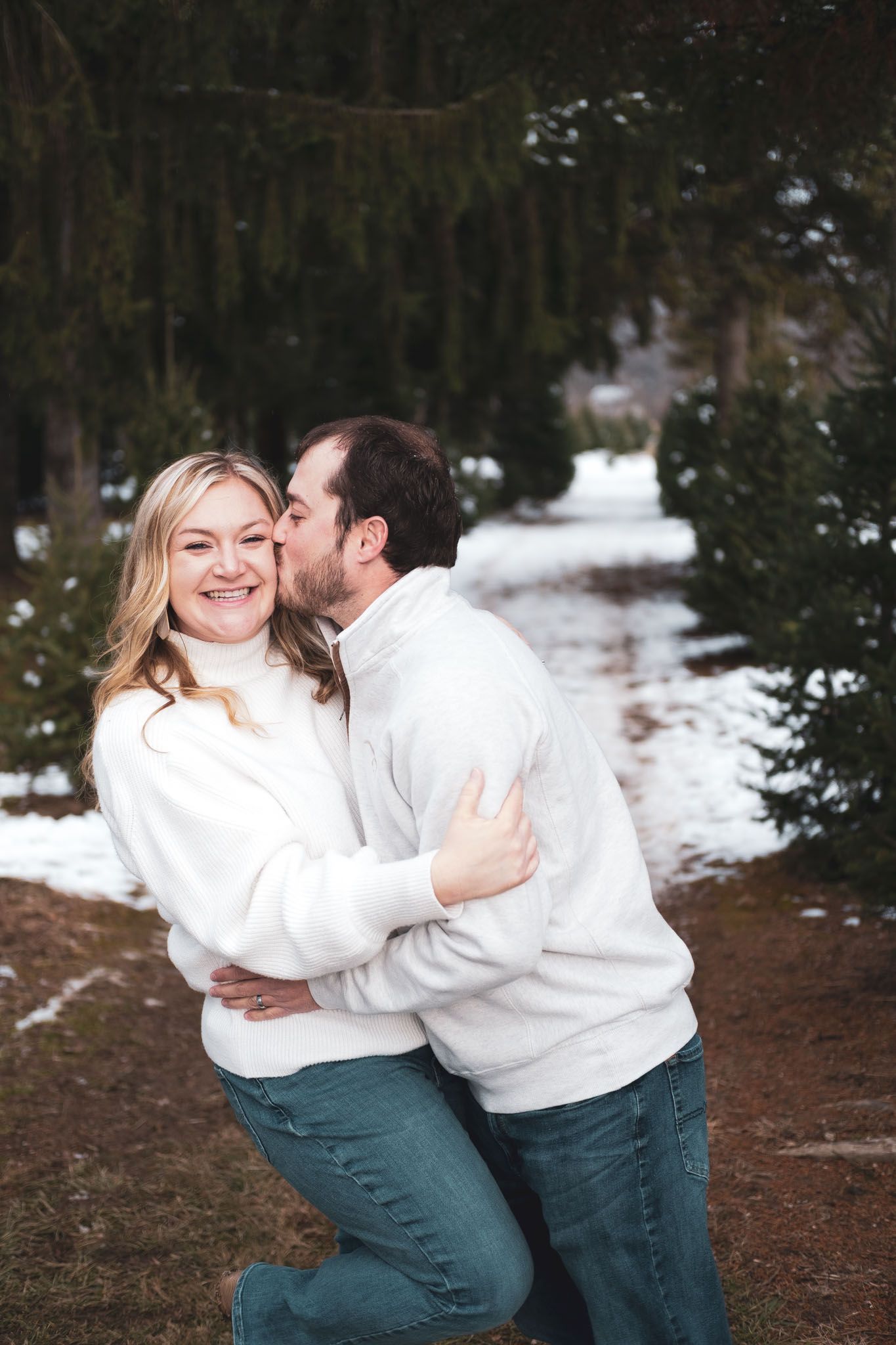 Smiling couple in white sweaters embracing outdoors on a snowy path under evergreen trees